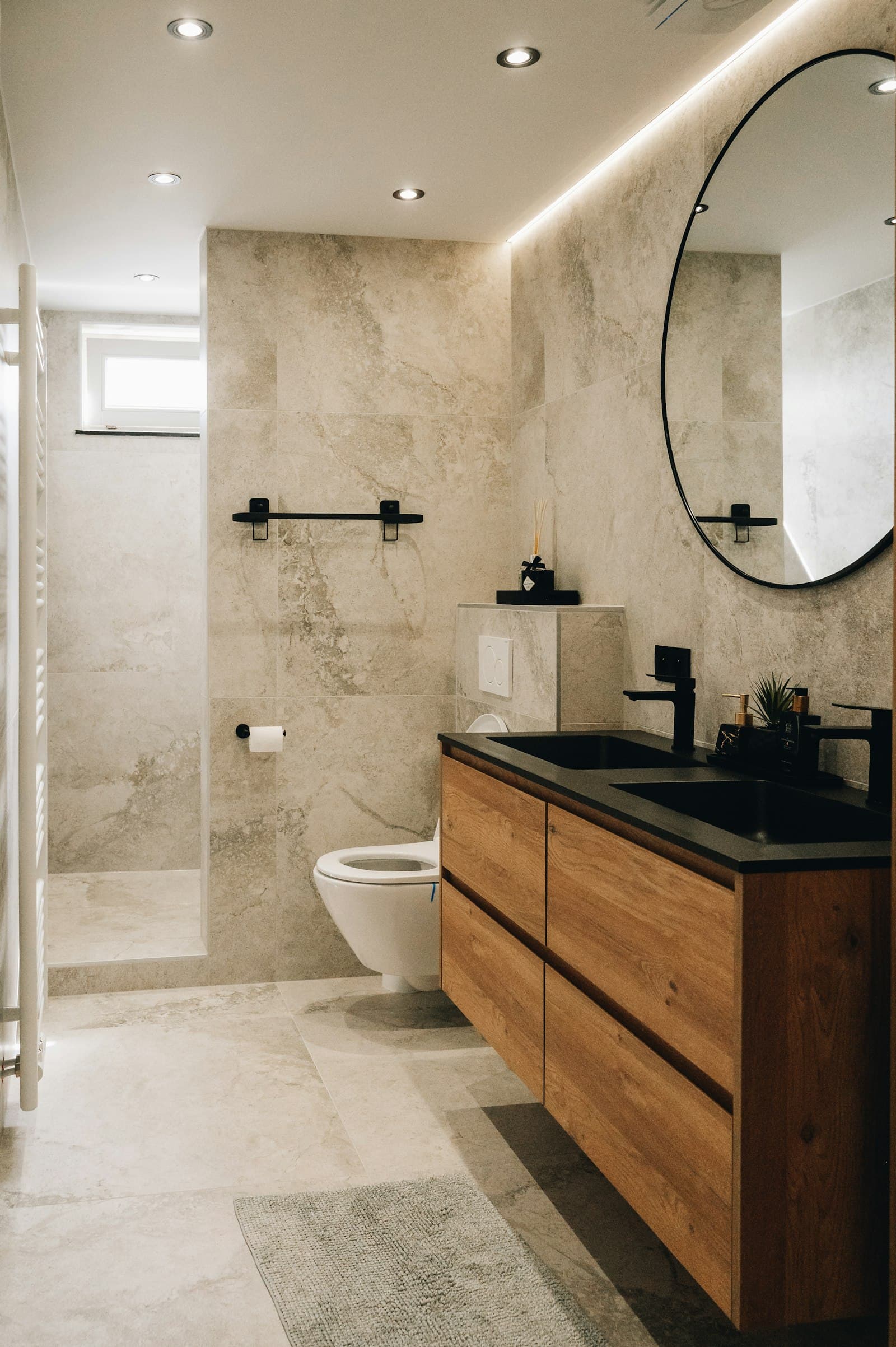 Bagno con rivestimento effetto pietra, mobile in rovere e specchio rotondo retroilluminato.